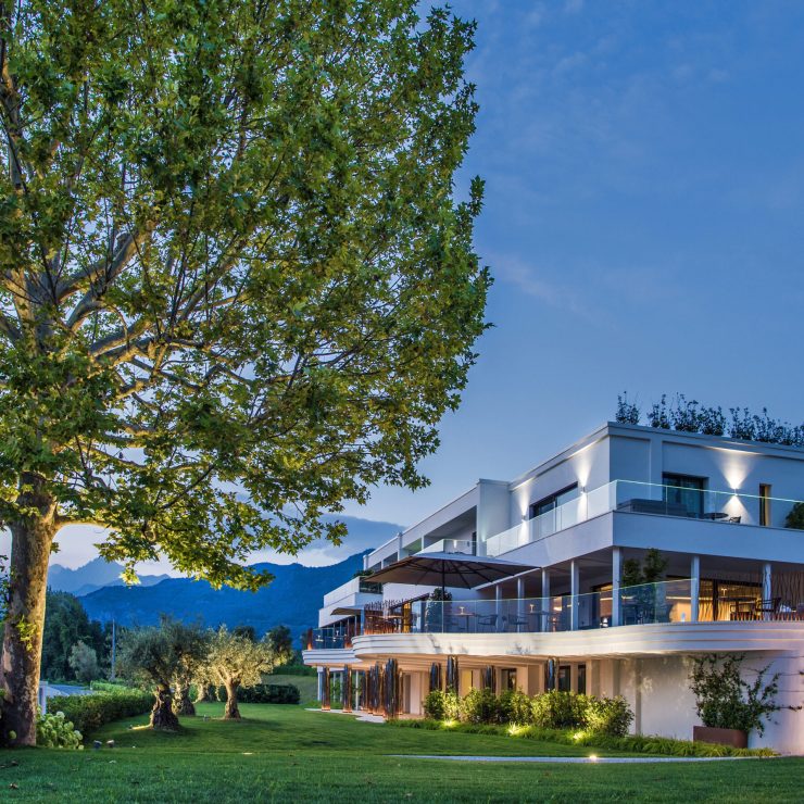 Esterno all'imbrunire, il volume bianco affacciato sull'acqua, il grande albero in primo piano. È l'unica foto che dà insieme architettura e contesto: come apertura funziona meglio di qualsiasi interno.