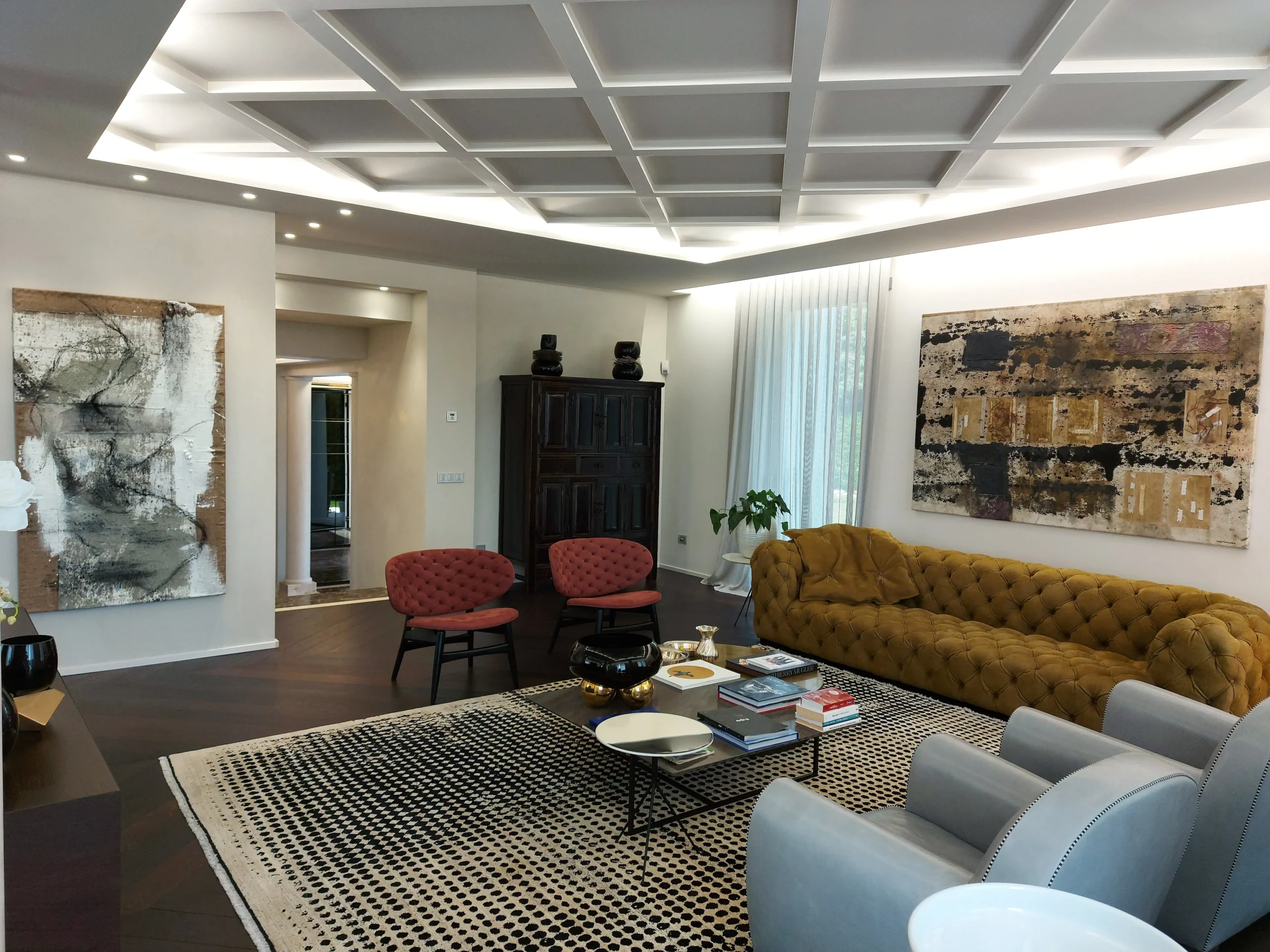 Salone con soffitto a cassettoni, divano capitonn&eacute; senape e grandi tele materiche