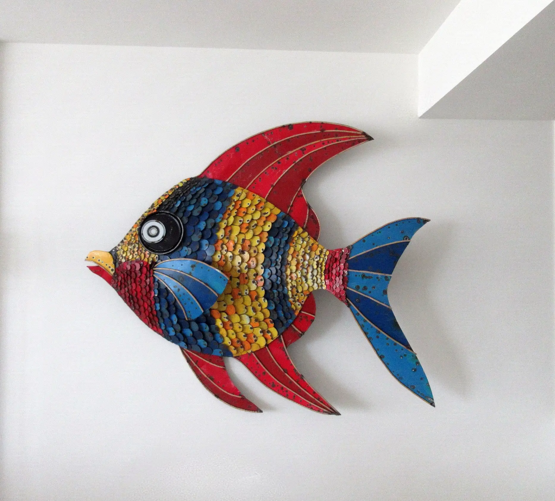 Scultura in metallo colorato a forma di pesce, appesa a una parete bianca